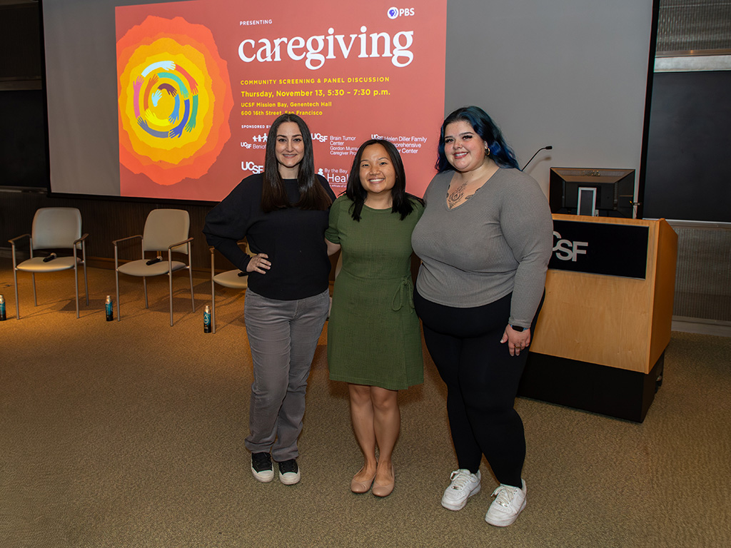 The three women standing together at the front of an auditorium. The screen behind them shows slide about the community screening of Caregiving.