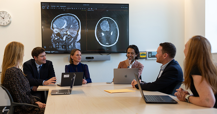 A group of people sitting around a conference table discussing a case. MRI scans of a meningioma are shown on the TV screens behind them.