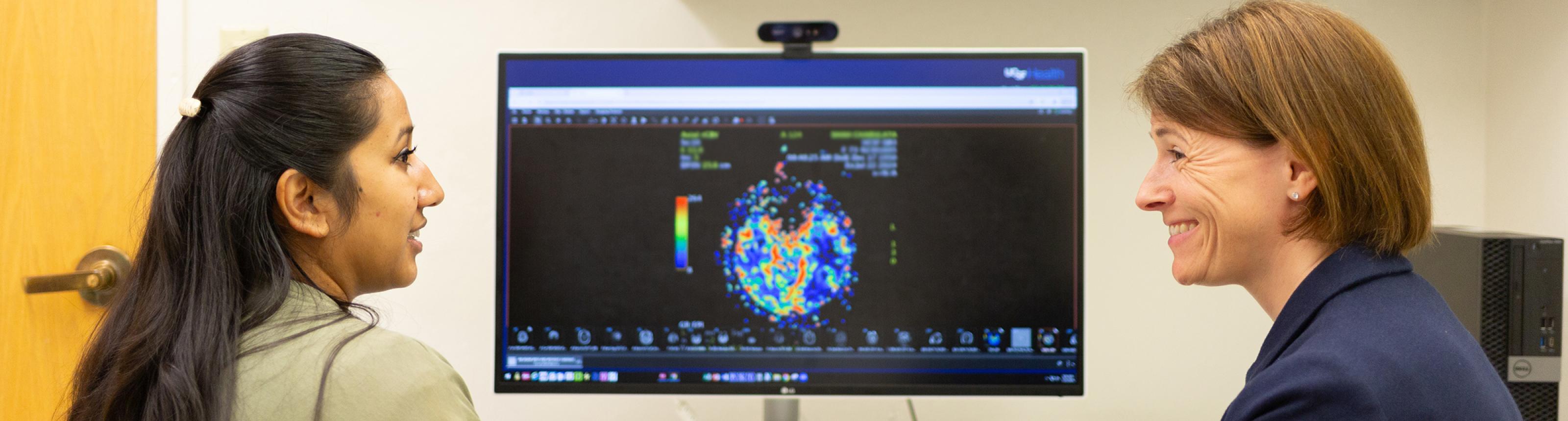 A physician smiling while talking to a patient. A color image of a brain tumor is visible on the computer screen behind them.